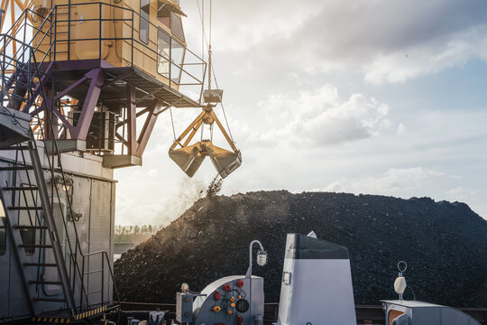 Heavy crane loads coal onto a vessel at the port on a clear day by the river. Loading coal onto a barge using a floating crane