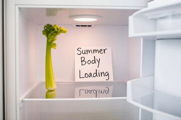 An empty refrigerator with a single stalk of celery next to a handwritten note that says Summer Body Loading