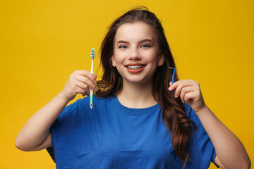 Cheerful teen girl with dental braces holds toothbrush and interdental brush. She wears blue t-shirt, stands on yellow background. Concept of oral hygiene, dental care, orthodontics, healthy lifestyle