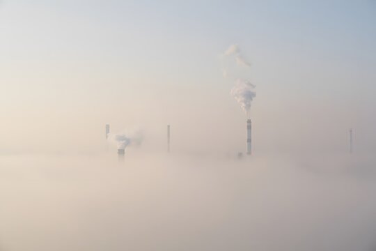 Factory chimney smoke pollutes the blue sky with white clouds as the sun rises over the industrial city landscape