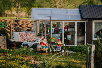 Colorful garden gnomes and cozy outdoor decor in front of a rustic greenhouse filled with vintage...