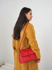 Woman in a mustard-colored coat with long dark hair, holding a vibrant red handbag over her...