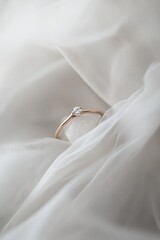 Delicate gold ring with a single, small, clear gemstone rests on a white stone, nestled in flowing white fabric.  Soft lighting enhances its subtle elegance