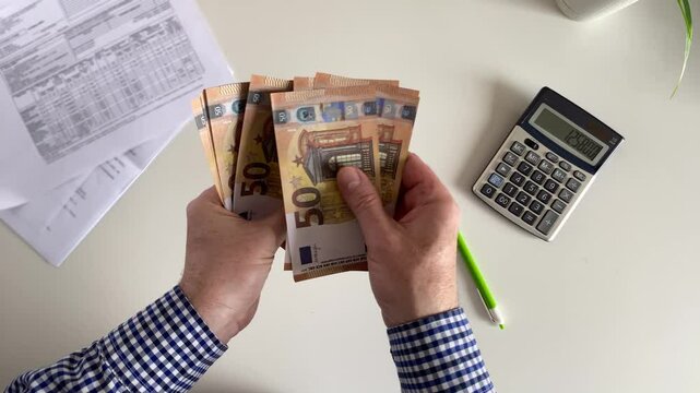 A man counts 50 euro paper bills against the background of utility bills and a calculator.