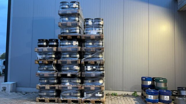 Stacks of metal beer kegs on wooden pallets outside a brewery, partially wrapped in plastic, with branded blue kegs to the right near a wall.
