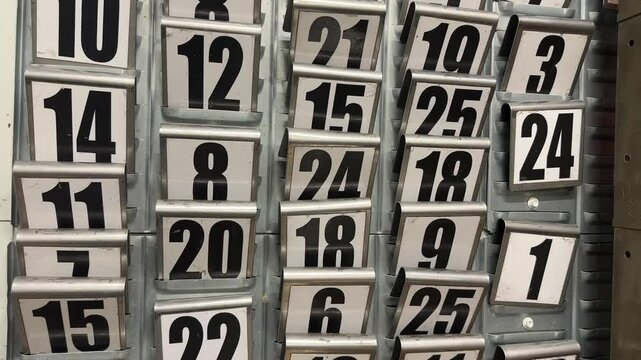 Wall-mounted numeric card slots with large black numbers, used for tracking employee shifts, locker assignments, or production queues in an industrial setting.

