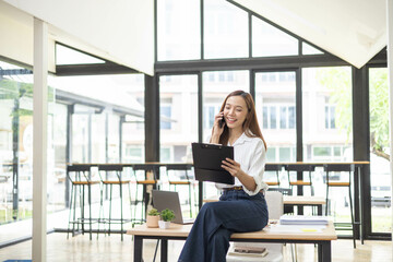 A woman is working at her desk with a laptop and a stack of documents. She is talking on the phone, looking at documents and seems focused on her work.

