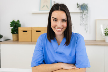 Smiling female nurse wearing blue uniform sitting at desk with arms crossed in bright doctor's...