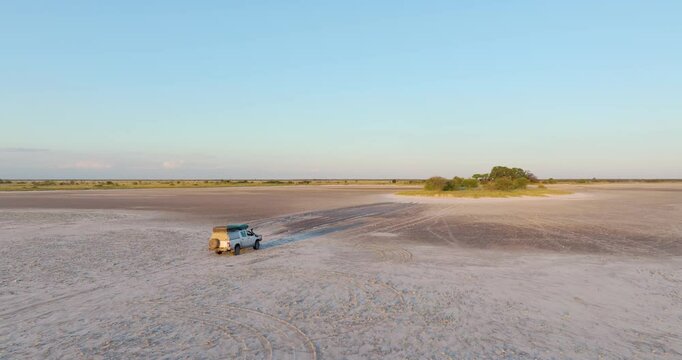 Drone footage of 4x4 off-road vehicle crossing salt pan toward tree-covered island in Nxai Pan, Botswana. Ideal for: travel content, safari promotion, overlanding, remote Africa landscapes.  