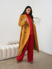 Standing woman in mustard coat and red outfit, holding a wine glass. Neutral-toned setting with...