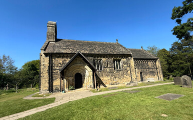 St John the Baptist church features arches and a sloping roof, set against a blue sky. A well-kept lawn surrounds the structure, adding to the serene atmosphere of the scene in, Adel, Leeds, UK