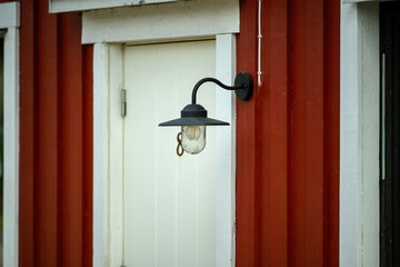 A black outdoor light hangs beside a white door on a red wooden wall. The rustic atmosphere highlights the charm of the structure in a serene environment