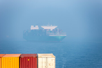 A large cargo ship laden with containers is partially obscured by fog, sailing on a calm sea. In...