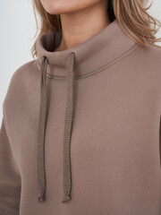 A close-up of a person wearing a taupe hoodie with a high collar and drawstrings. The focus is on...