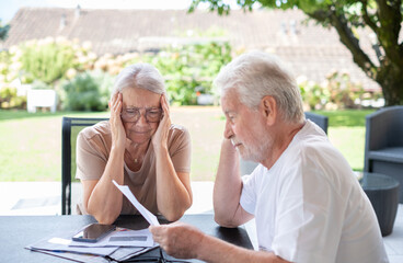Elderly couple sitting at table at home with worried expression while reading paper documents, paying bills, managing bank finances, calculating taxes, planning loans, debts, insurance and pensions.