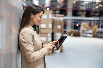 Woman manager in professional attire using tablet for inventory check, standing in a warehouse.