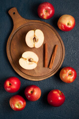 Fresh red apples, both whole and halved, arranged around a round wooden cutting board with two cinnamon sticks on a dark textured background. A cozy and warm autumn-inspired flat lay scene.