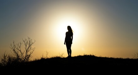 Fototapeta premium Silhouette of a woman standing on a hill against a bright sunset sky, capturing a moment of peace and reflection