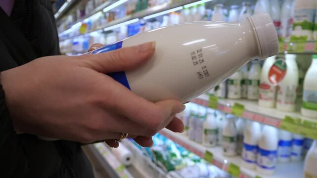 Close-up of female hands holding a bottle of milk in a store and reading the label describing the ingredients and expiration date. Selection of dairy products, fat content, dairy products, natural pro