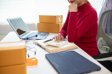 Asian businesswoman in red shirt is checking product orders and calculating sufficient stock to deliver to customers according to orders entered into online shopping application.Copy Space for text