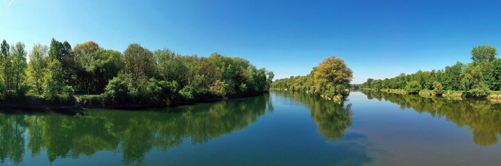 Die Lechmündung in die Donau zeigt eine friedliche Flusslandschaft mit üppiger Vegetation und klarem Wasser. Die Spiegelung der Bäume im Wasser und der blaue Himmel schaffen eine idyllische Atmosphäre