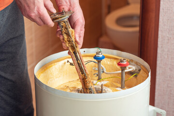 A worker holds a heating element of an electric boiler covered in rust and mineral deposits. Boiler maintenance and repair