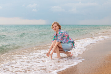 Woman in floral shirt and denim shorts squatting playing in shallow waves on sandy beach. Summer vacation at sea