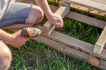 A man uses a drill to assemble patio furniture from a wooden pallet on the grass in a garden