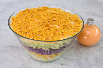Seven Layer Salad in a clear glass bowl with layers of shredded lettuce, corn, red cabbage, green cabbage, creamy sauce bottle, and a crunchy crushed chips side view of classic and colorful salad