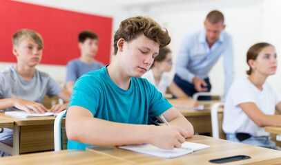 Diligent teenage high school student studying with classmates, making notes of teacher lecture