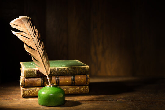 Feather and ink bottle with old books on dirty wooden table. Feather for calligraphy, old educational, antique leather-bound book covers with golden ornament.