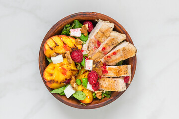 Grilled chicken breast salad with peaches, feta cheese, raspberries, walnuts and mixed greens in a bowl on white marble background. Top view. Healthy diet protein food. Summer dinner