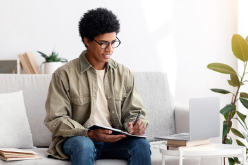 Distance education concept. Focused black student studying remotely on laptop computer at home. Busy teenage guy taking notes during web conference, online lesson or webinar indoors