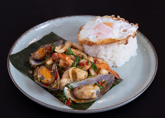 A plate of Thai traditional spicy stir fried with basilic variety of mixed seafood. White rice topped with crispy mirror egg, black isolated background.