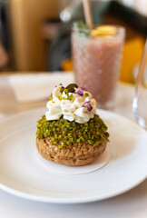 A French cream puff crack bun (Choux au Craquelin)topped with rose cream and pistachio with its grains topped with rose petal. Soft drink in background. It's a dessert base of savory choux dough bun.