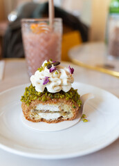 A cut French cream puff crack bun (Choux au Craquelin) topped with rose cream and pistachio with its grains and rose petal. Soft drink in background. It's a dessert base of savory choux dough bun.