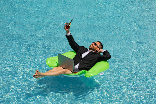 Excited business man floating in suit drink summer cocktail and using laptop in pool. Travel business. Office employee using laptop in pool on summer day. Successful businessman dreams in summer pool.