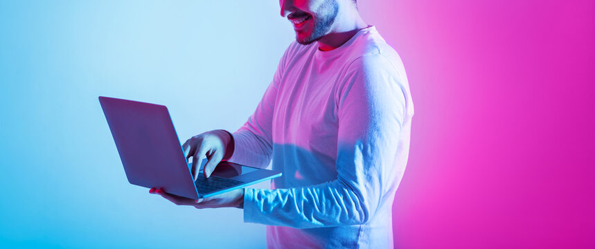 Freelance and work remotely, online game and correspondence. Contented millennial guy use laptop and holds in hands on pink and blue background, panorama, copy space