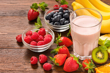 Top view of glass with summer strawberry smoothie surrounded by fresh berries bananas kiwi orange and mint leaves on rustic wooden table with copy space
