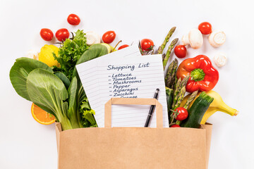 Top view of paper shopping bag with fresh vegetables and grocery list on white background. Healthy...