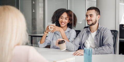 Obraz premium Coffee break in modern office during coronavirus outbreak and social distancing. Multiracial colleagues with cups of hot drink talk through protective glass at table with antiseptic, empty space