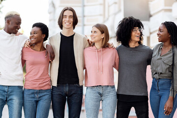 Obraz premium Friendship, community, team concept. Multiracial group of happy millennial men and women posing on the street, standing in a row, embracing and cheerfully smiling at camera, full length shot, panorama