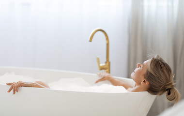 Relaxed blonde lady enjoying hot foamy aromatic bath at home, lying in tub full of water with closed eyes, side view, closeup photo, copy space. Home spa, body care concept