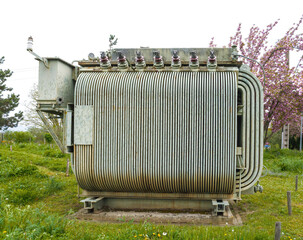 Large outdoor electrical transformer with ribbed cooling fins and high-voltage insulators stands on a concrete base in a grassy field surrounded by flowering trees and utility poles.