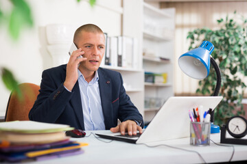 Concentrated businessman solving business issues by phone, sitting in office with laptop