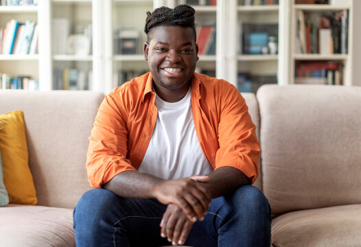 Portrait of happy handsome young black man in casual outfit with braids sitting on couch at home, smiling at camera, living room interior, copy space. Millennials lifestyles concept