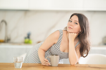 Young positive woman housewife leaning on table with cup in kitchen at home