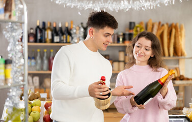 Happy carefree young couple exploring wine options in local grocery store decorated with shiny tinsel for Christmas, selecting drinks for holiday party..