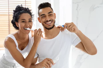 Acrylglasbilder Zahnärzte Cheerful young arab couple brushing teeth together in bathroom, happy middle eastern spouses doing morning oral hygiene and smiling to their reflection in mirror, joyful lovers bonding at home  © Prostock-studio