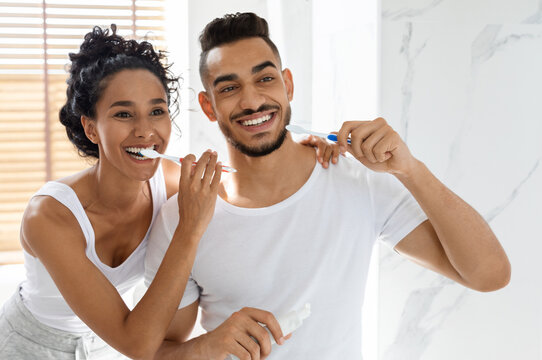 Cheerful young arab couple brushing teeth together in bathroom, happy middle eastern spouses doing morning oral hygiene and smiling to their reflection in mirror, joyful lovers bonding at home